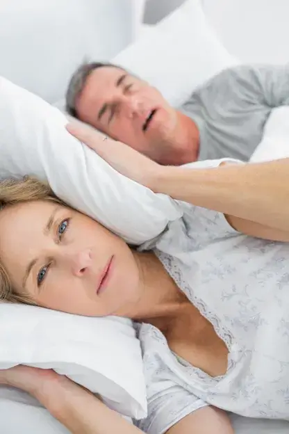 Frau hält sich mit einem Kissen die Ohren zu, da ihr Mann schnarcht